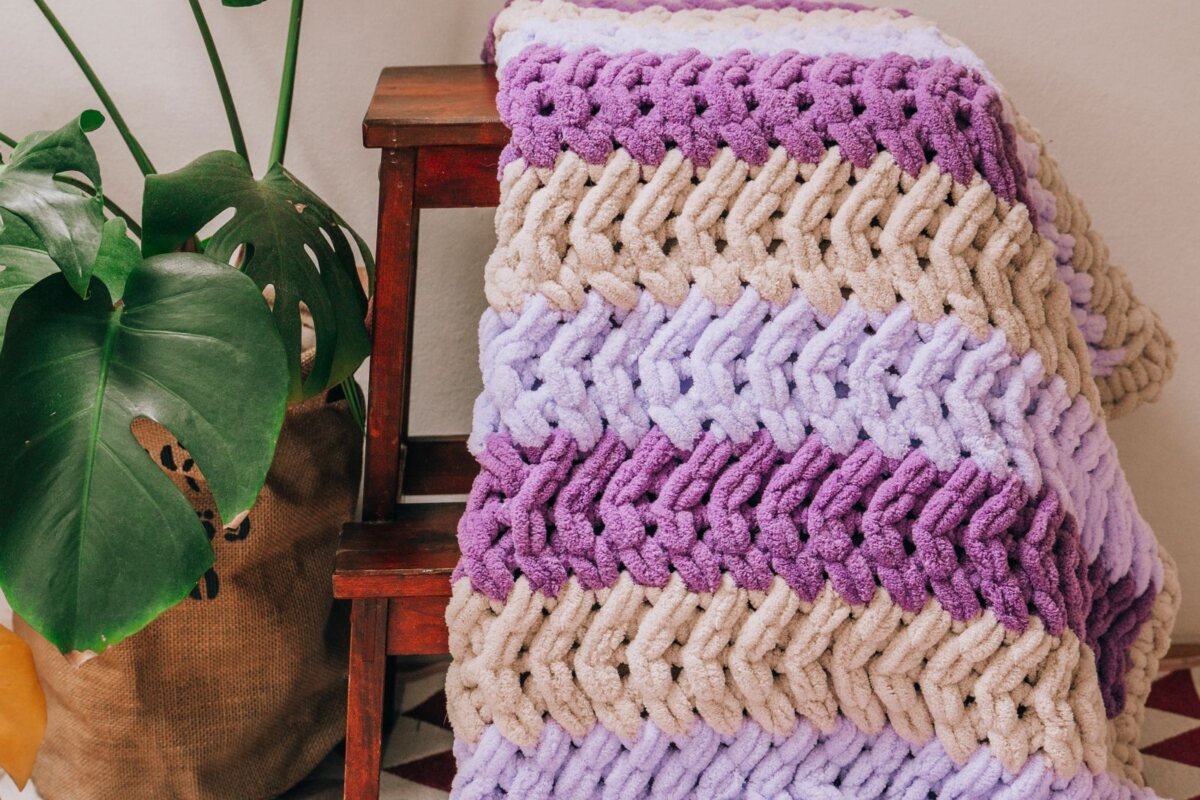 A chunky chenille crochet blanket in lilac and cream stripes folded over a wooden stool