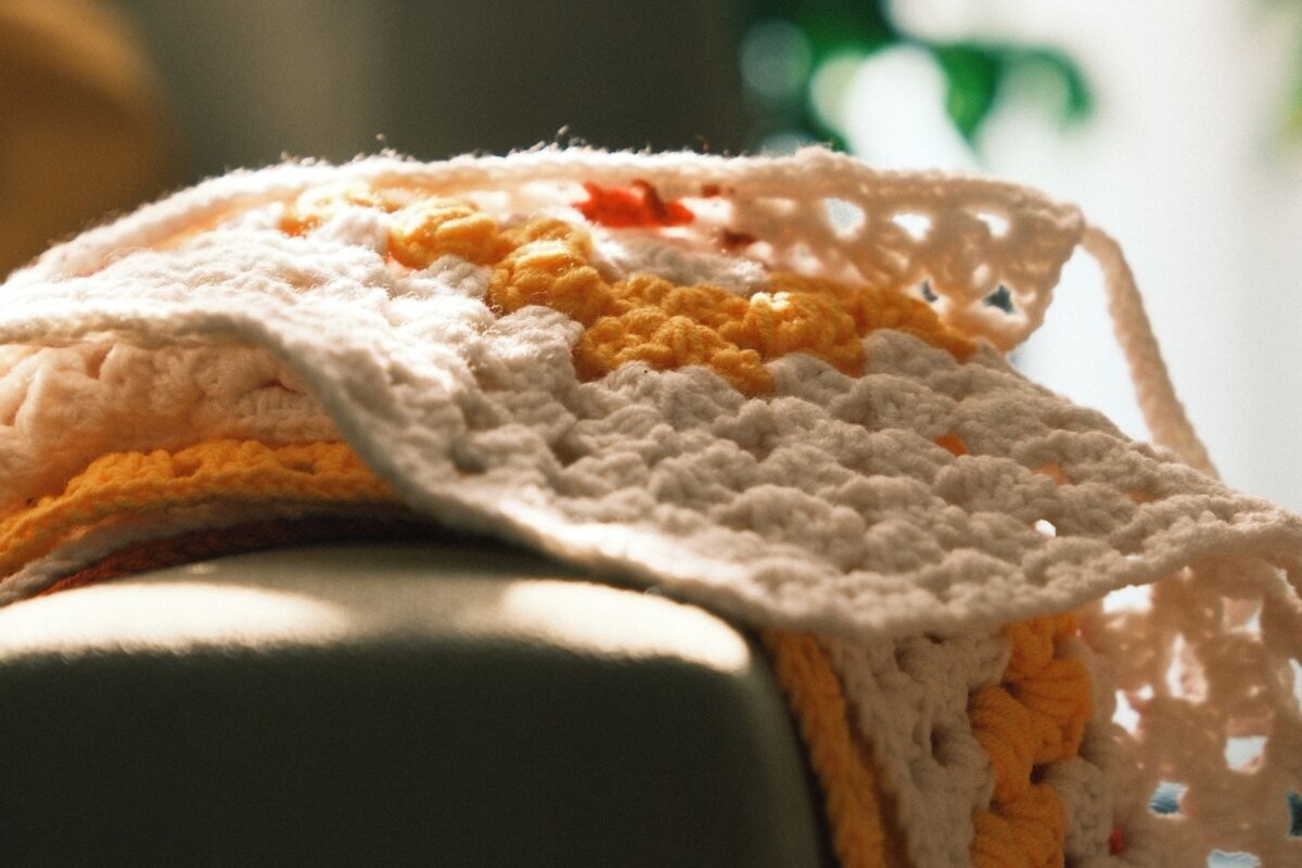 A cream and marigold crochet blanket folded over the arm of a sofa in warm sunlight