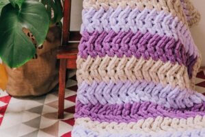 A crochet blanket worked in one repeating row pattern, folded over a wooden stool