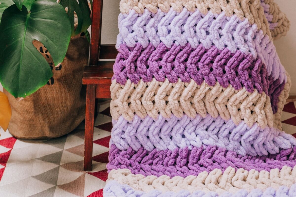 A crochet blanket worked in one repeating row pattern, folded over a wooden stool