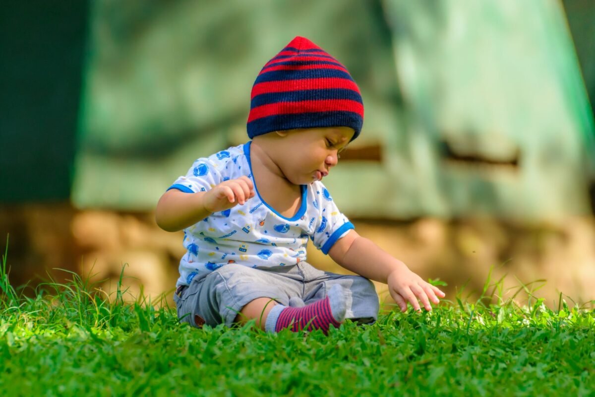 A baby in a striped beanie sitting on grass outdoors