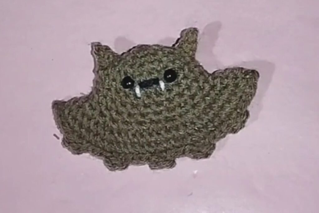finished crochet bat keychain in tan yarn with black eyes and white fangs lying on a pink surface