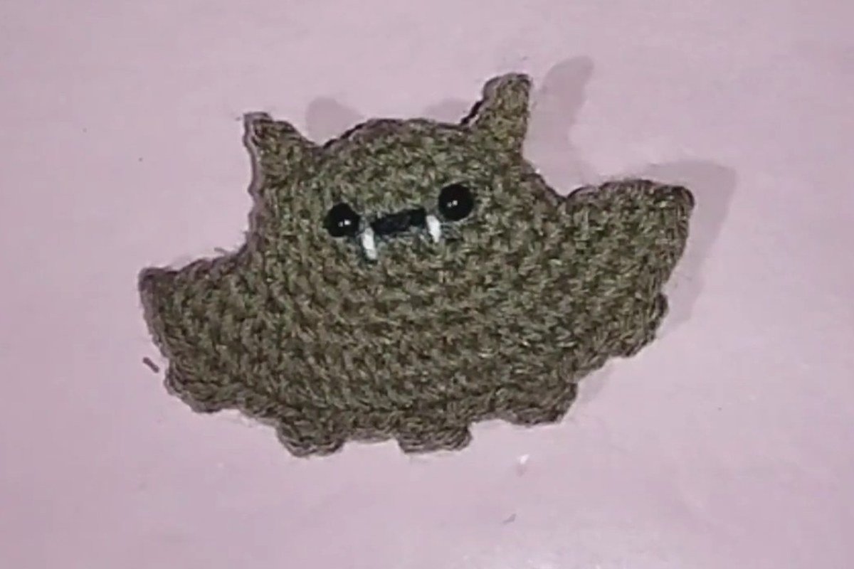 finished crochet bat keychain in tan yarn with black eyes and white fangs lying on a pink surface