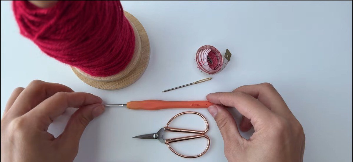 materials for a free crochet fingerless gloves pattern: red yarn, a 2.5 mm hook, scissors, a tape measure and a yarn needle