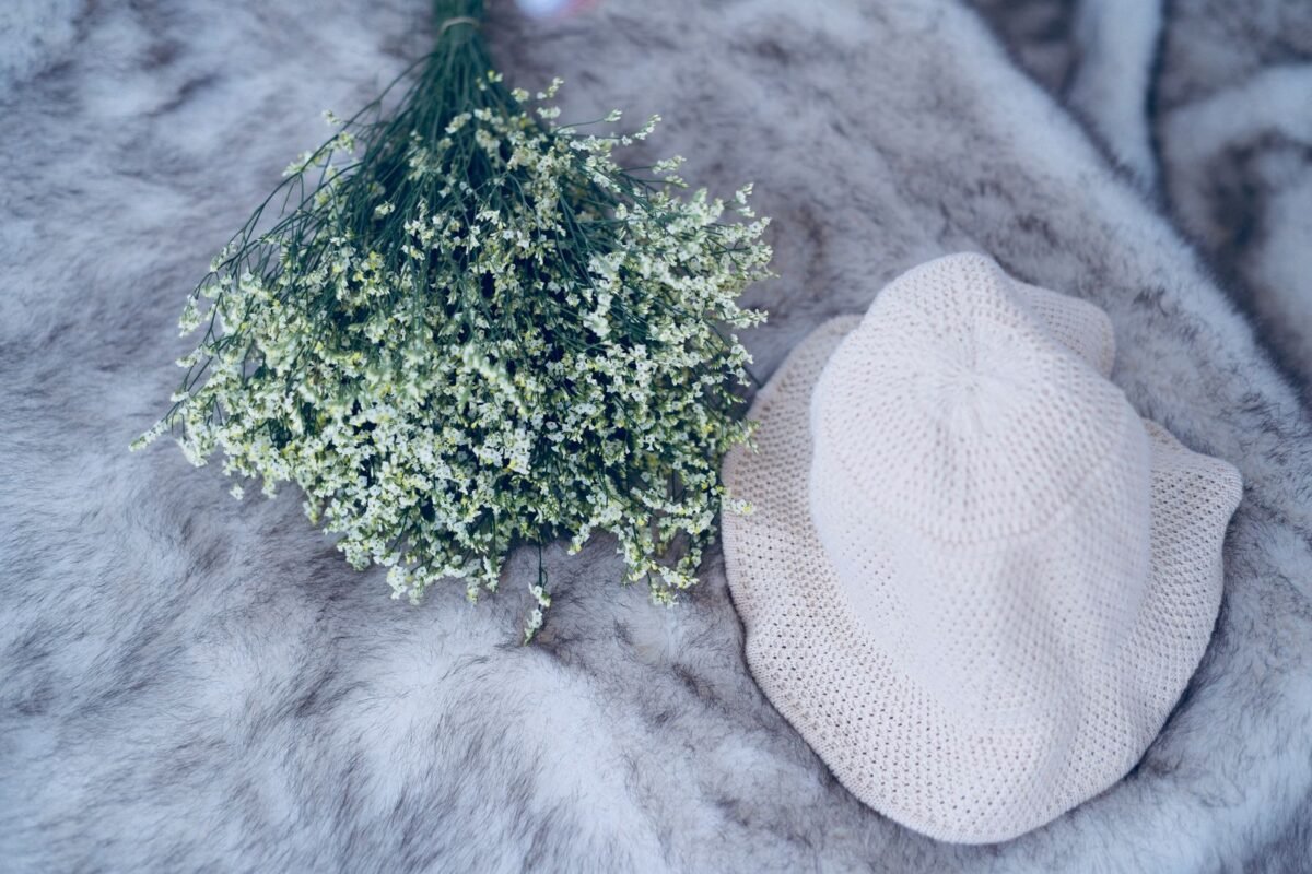 A small white crochet baby hat laid flat on a soft grey rug beside a bunch of dried flowers